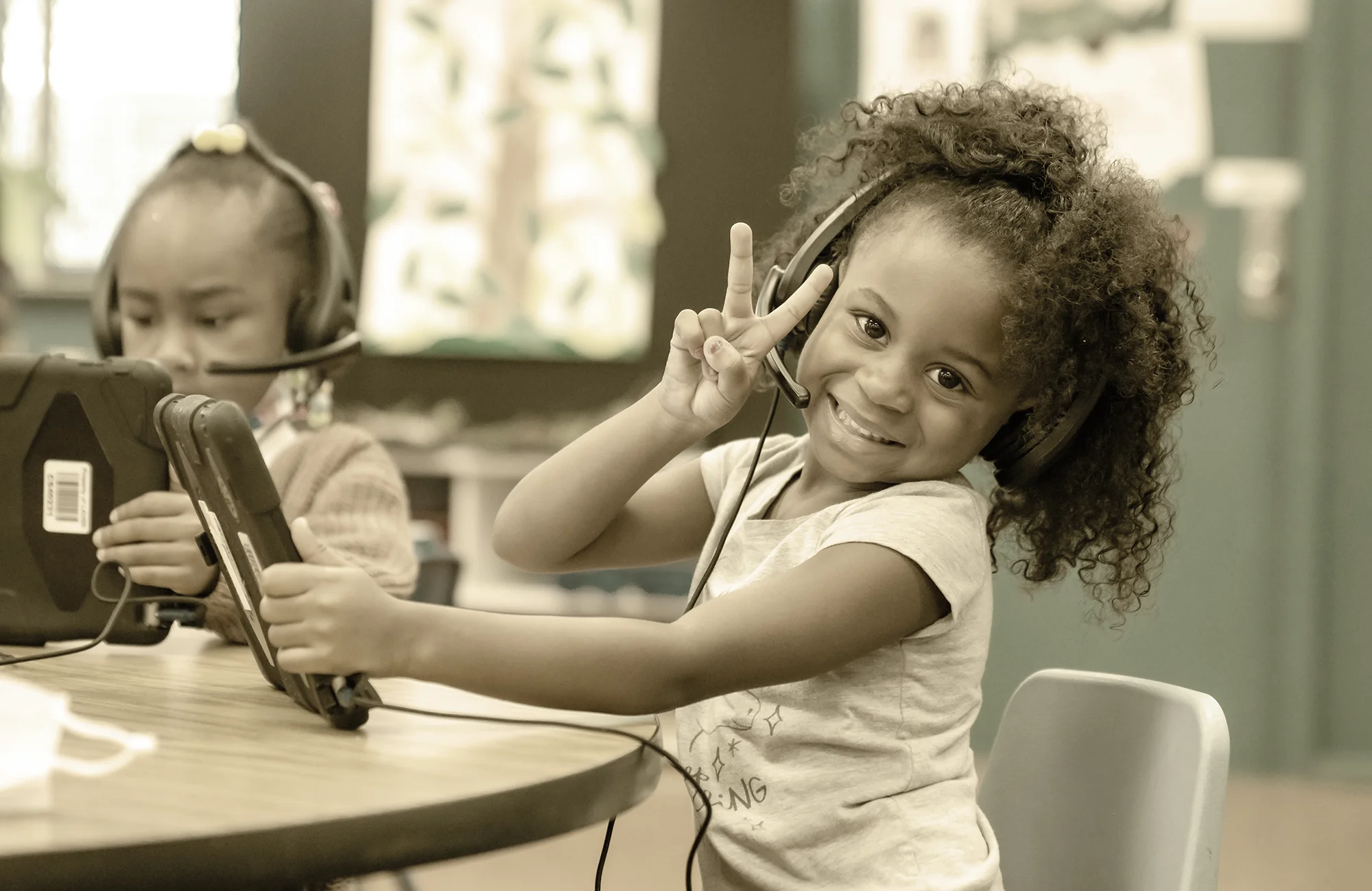 Happy young student wearing headphones and holding a tablet while giving a peace sign in a classroom digital learning center.