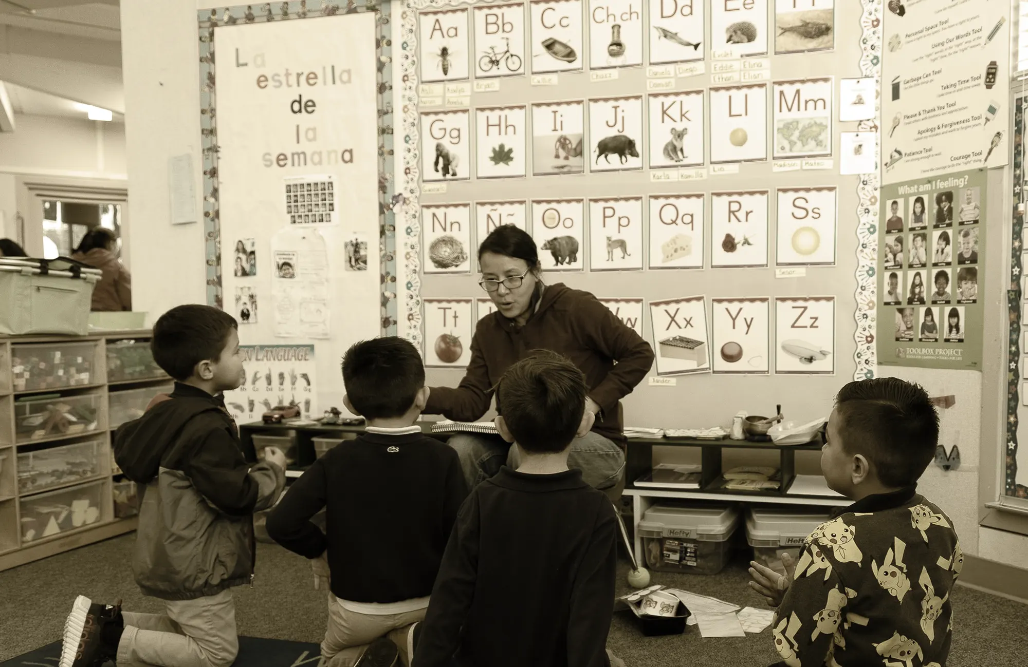 Elementary school teacher sitting with a small group of young students in a classroom near a wall displaying a phonics and alphabet chart.