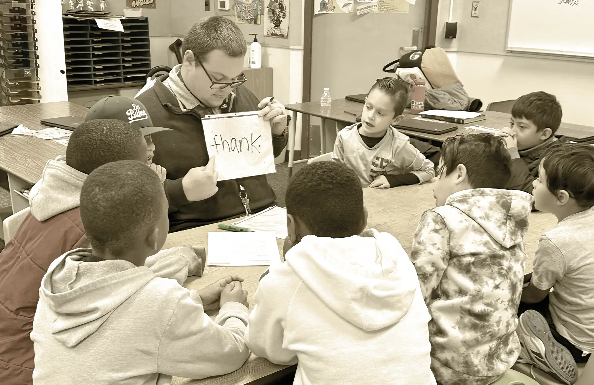 Teacher leading a small group of elementary students in a reading or phonics lesson, holding up a card with the word 'thank' written on it.