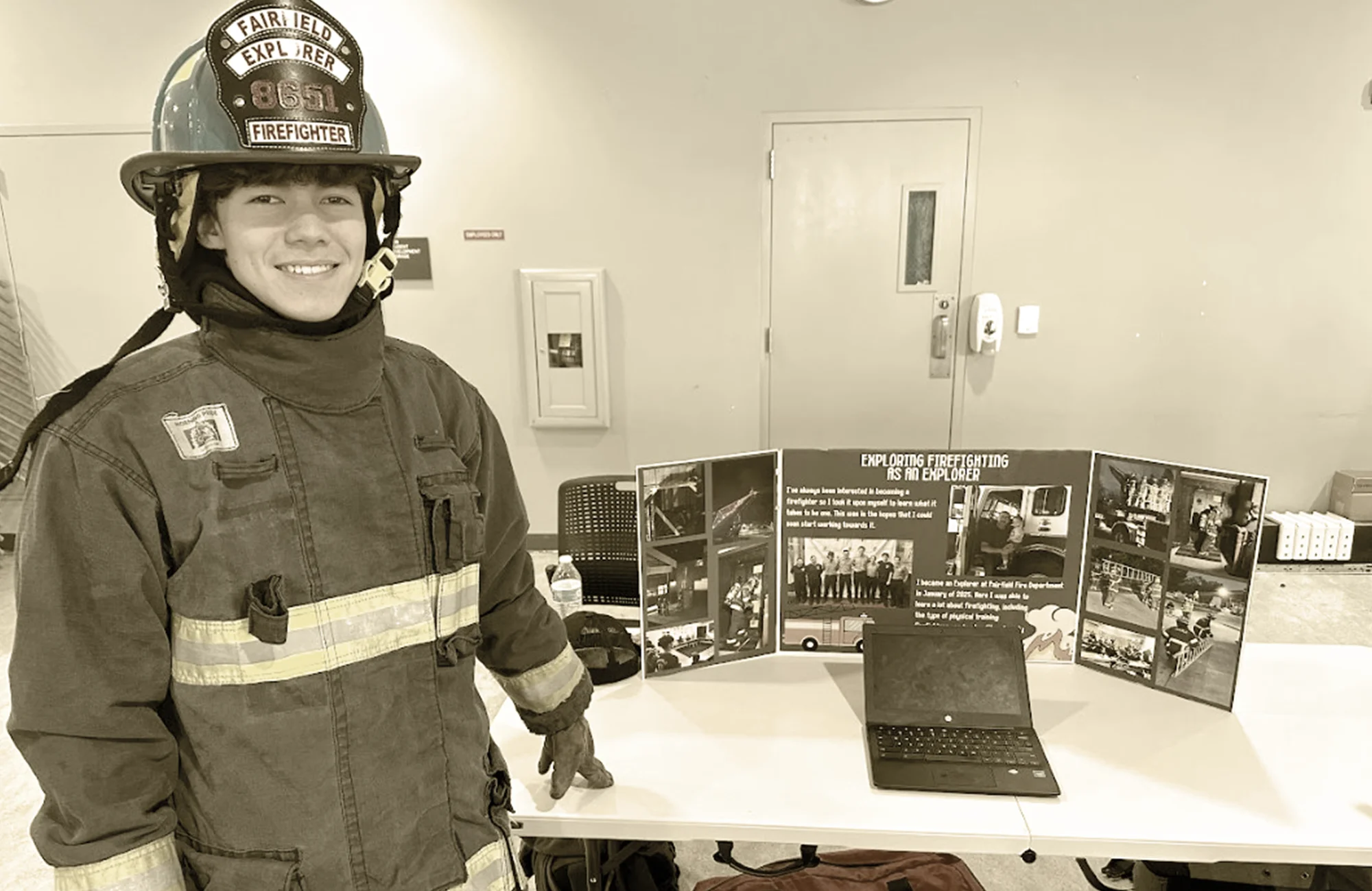 Young man in a fire helmet and bunker coat standing next to a display board and a laptop at a career fair.