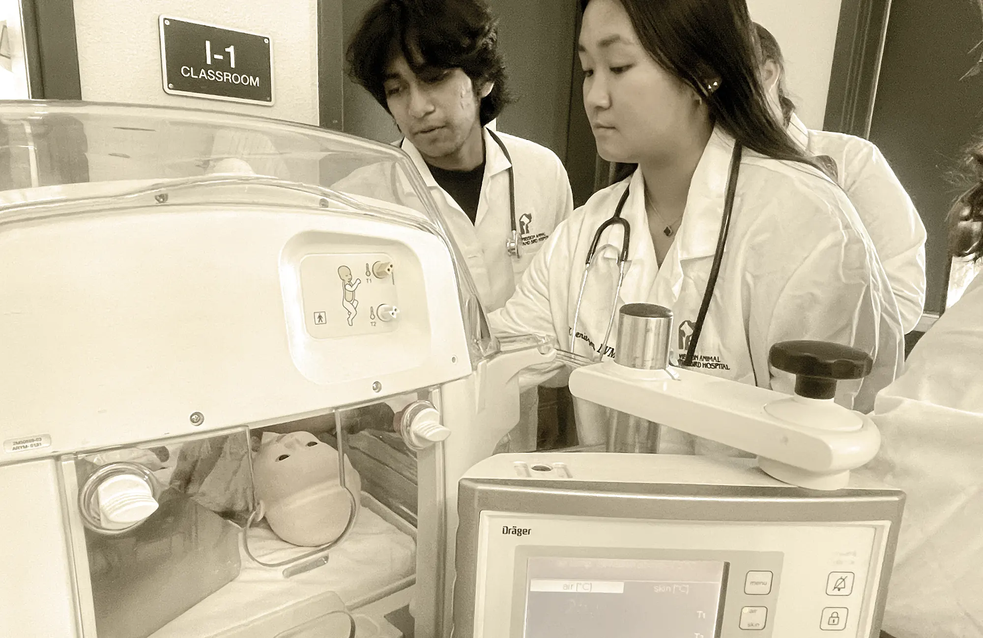 Career Technical Education (CTE) students in lab coats and stethoscopes training with a baby mannequin in a neonatal incubator.