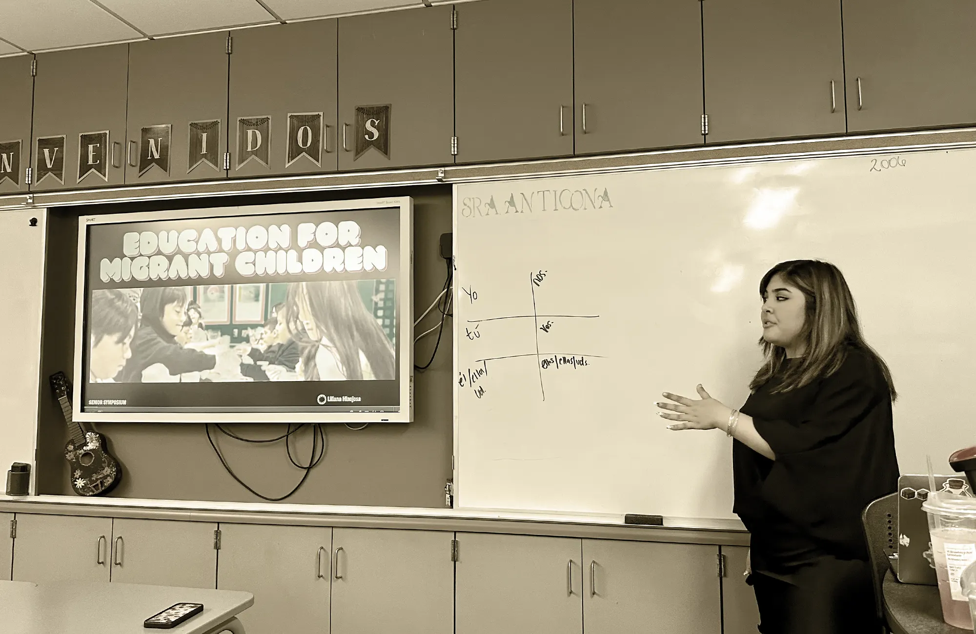 Classroom scene of a teacher presenting on a whiteboard next to a monitor displaying the title 'Education for Migrant Children'.