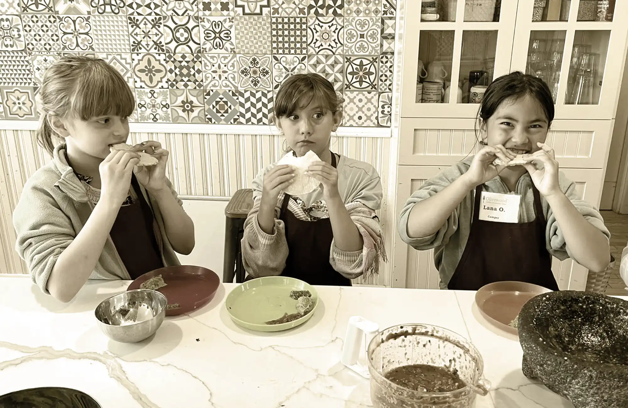 Girls at a table tasting food in a brightly lit kitchen setting with patterned walls.