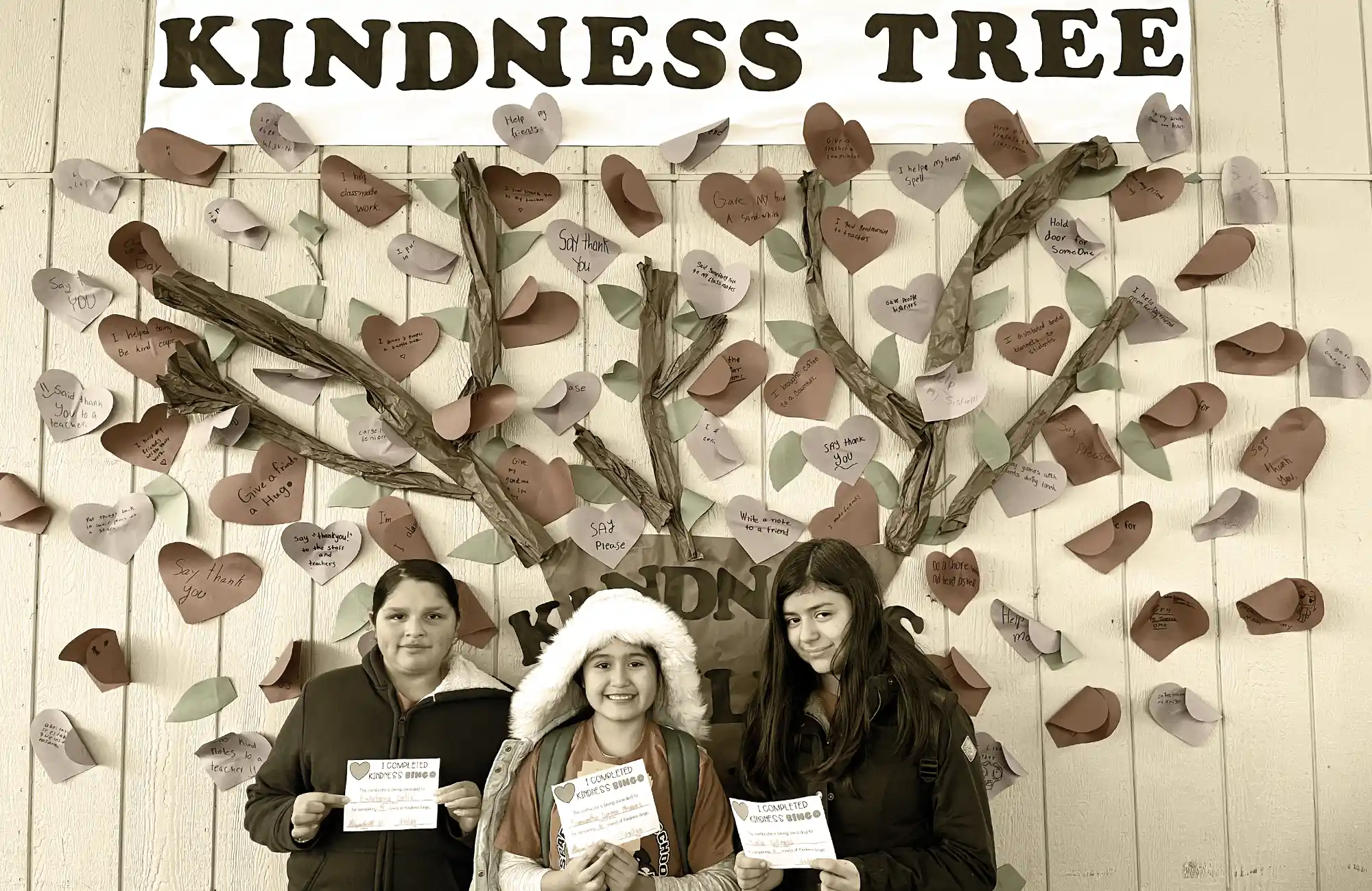 Three students holding kindness pledges in front of a school display titled 'Kindness Tree,' covered in paper hearts with kind messages.