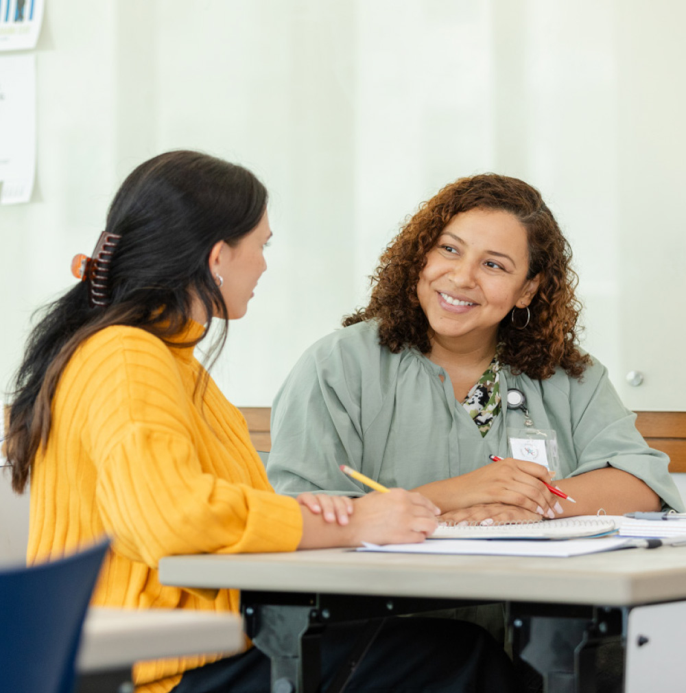 Two smiling women, one in a yellow sweater and one in a light green top with an ID badge, sit across a table talking and writing notes.