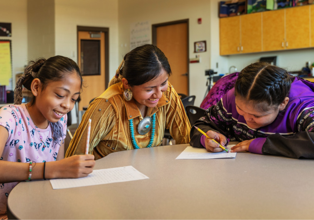A smiling woman mentoring two young girls writing at a round table in a classroom.