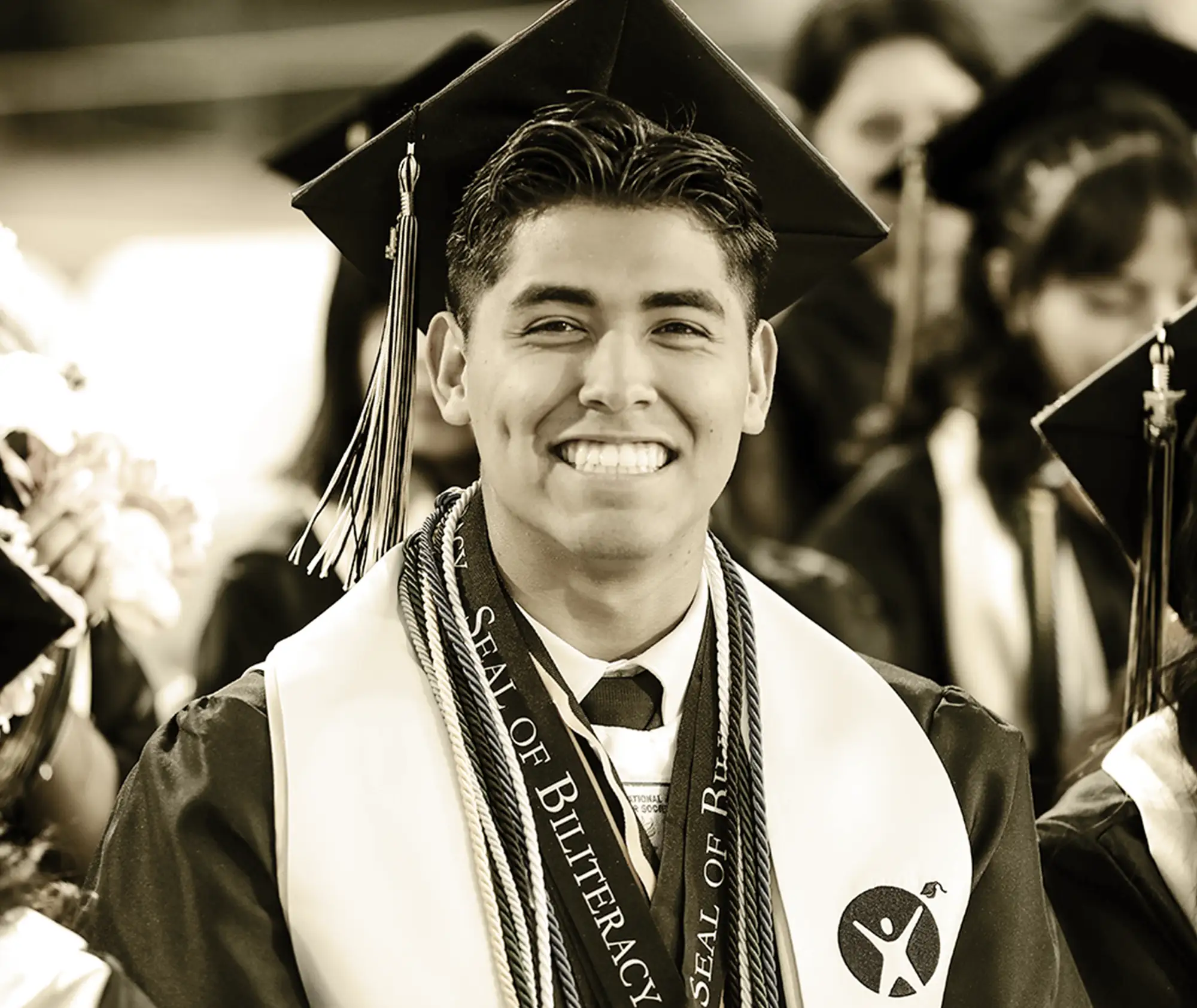 a smiling male graduate wearing a cap, gown, and a sash labeled "Seal of Biliteracy."