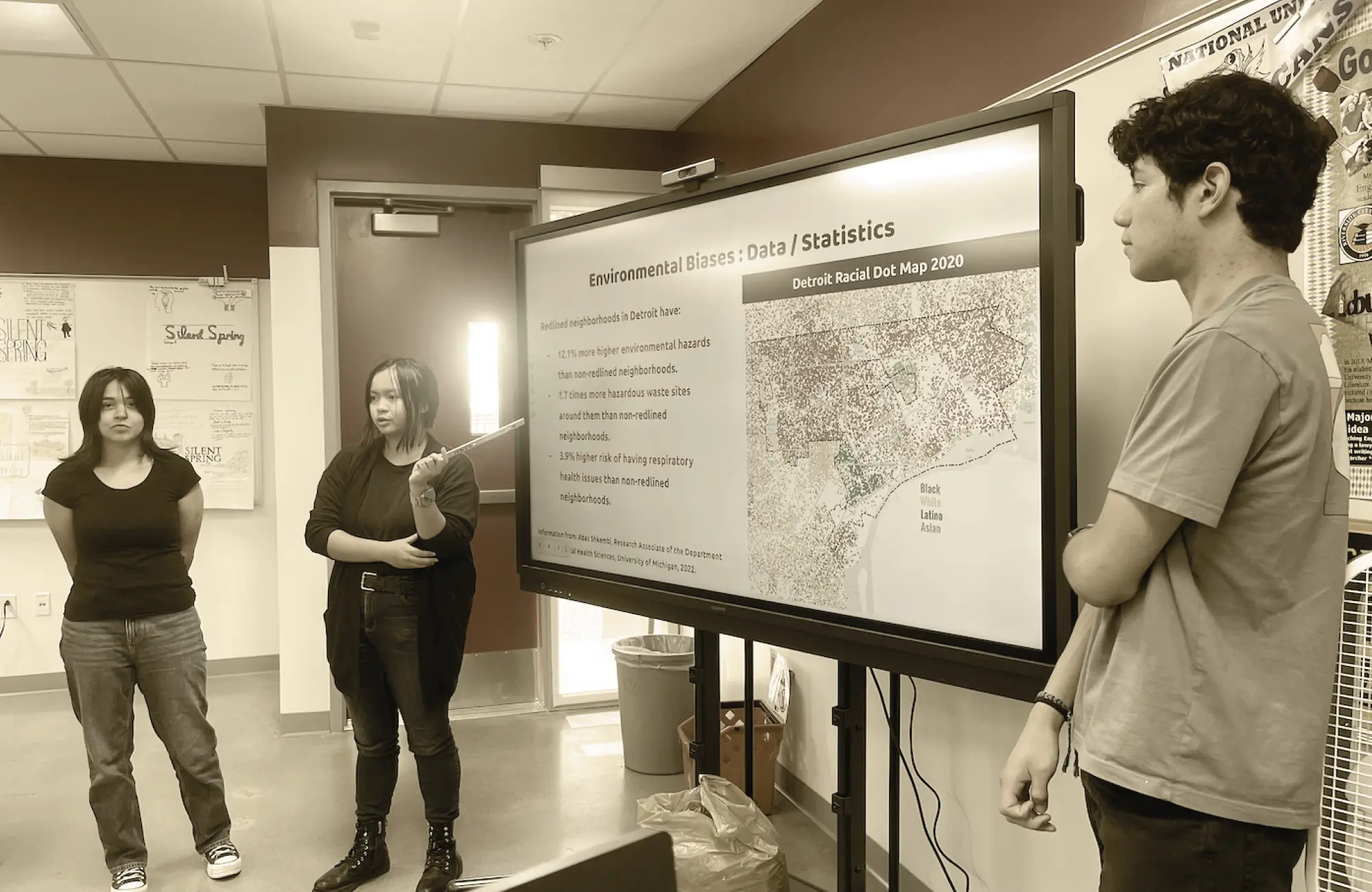 Three high school students presenting a project on environmental bias. One student points at a screen displaying a Detroit racial dot map and text about disparities in redlined neighborhoods.