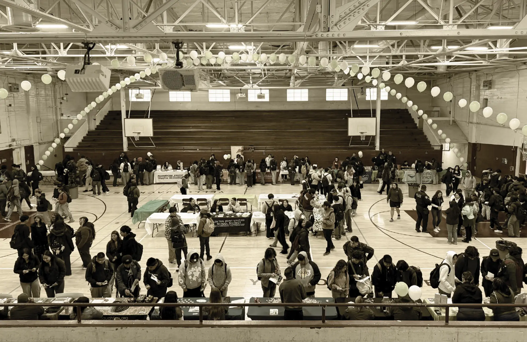 Wide shot of a large number of students and adults attending a resource fair inside a high school gymnasium. White balloons arch over the crowd and multiple resource tables are set up on the floor.