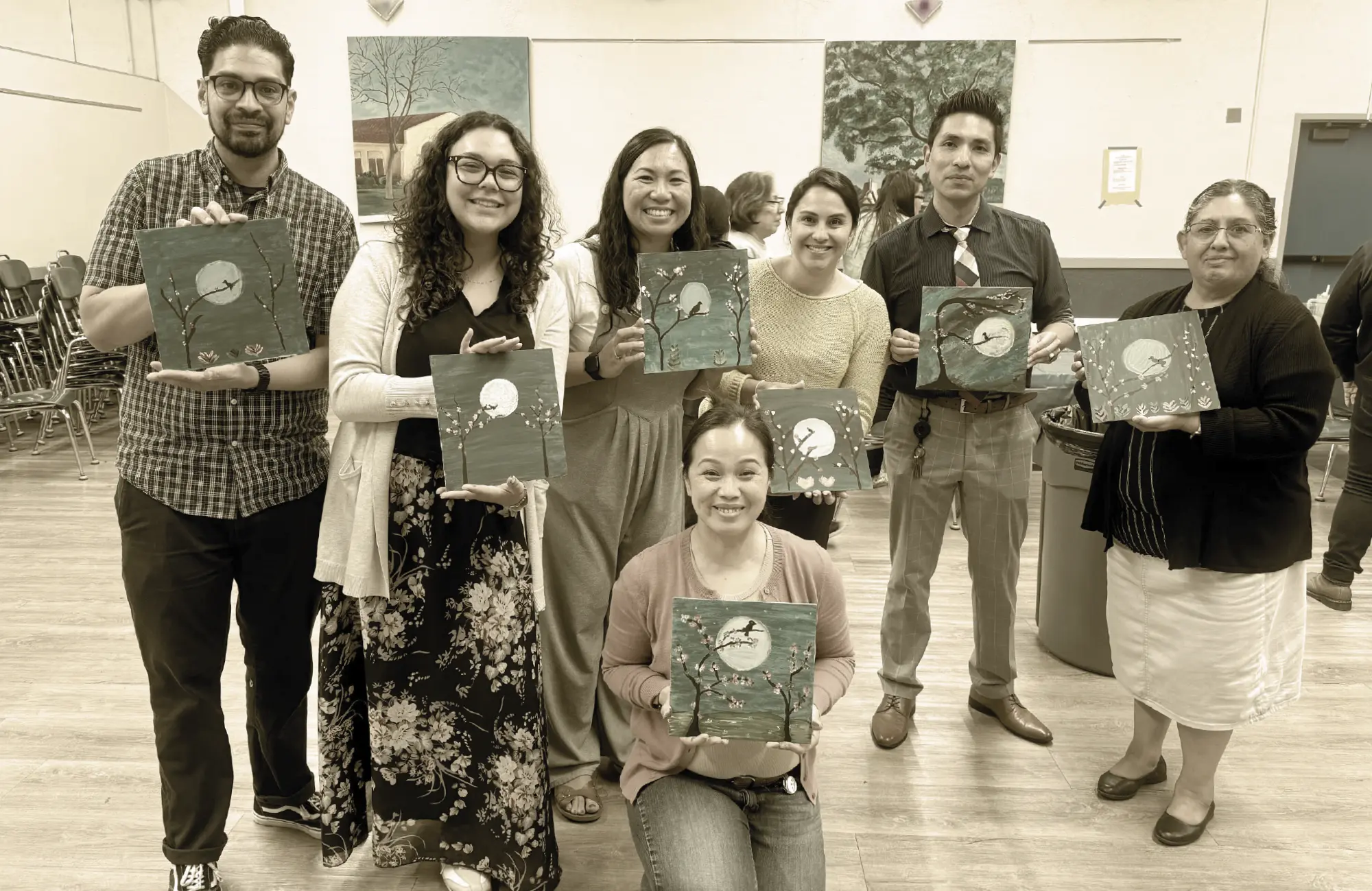 Seven adults, smiling and standing in a school setting, proudly hold up small painted canvases featuring a tranquil scene of a bird on a branch under a full moon. They represent the Wellness Committee.
