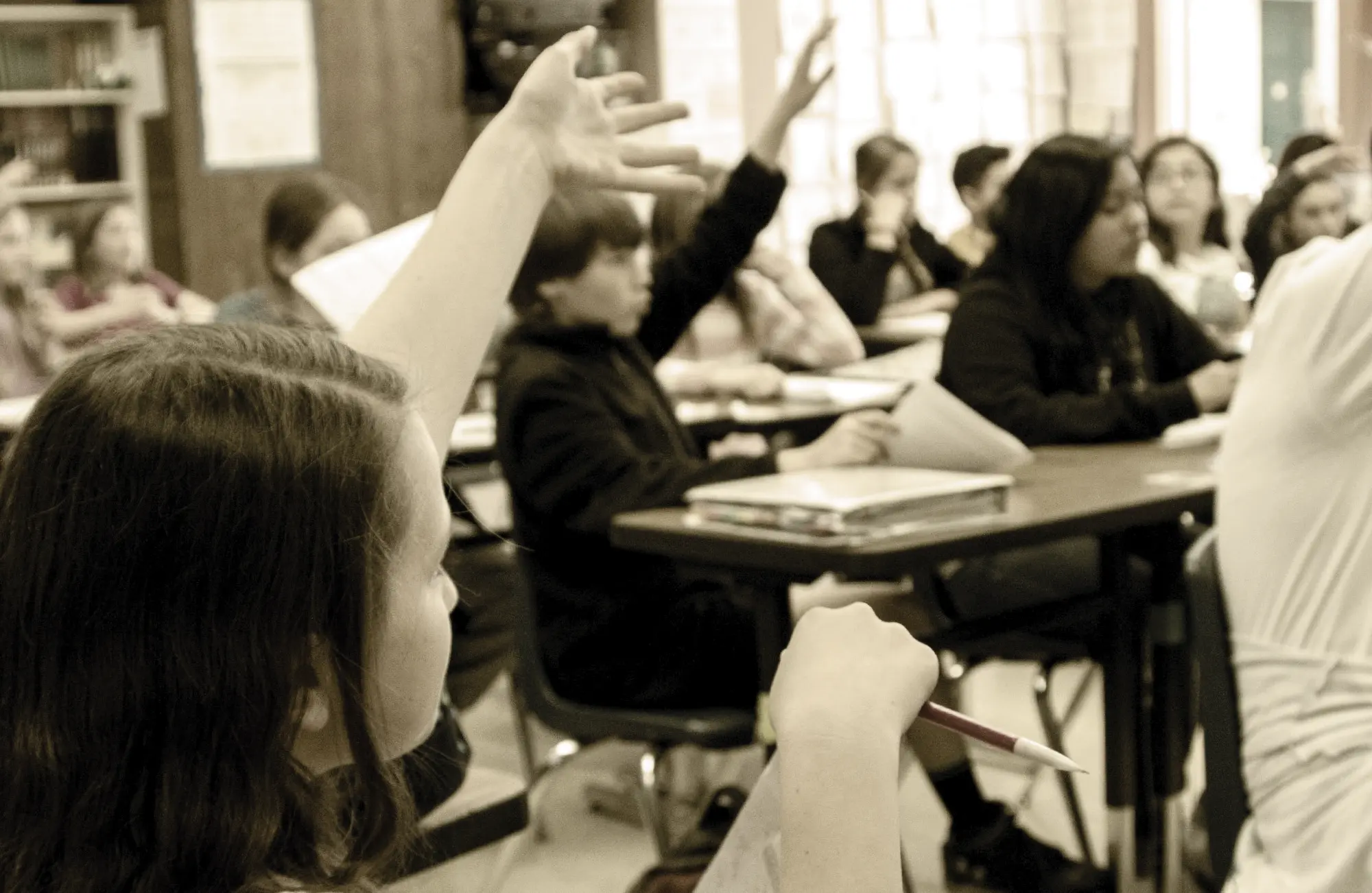 Close-up, over-the-shoulder view of a student in a classroom raising their hand to answer a question. Other students are seated at desks in the background, with several others also raising their hands.