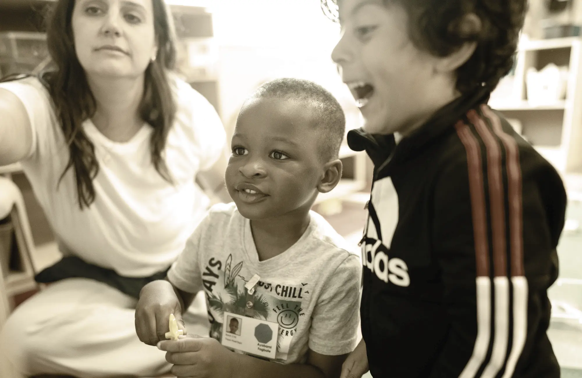 Close-up of two young, happy preschool boys sitting together with a teacher visible in the background. The image reflects the integrated setting of an inclusive preschool classroom.