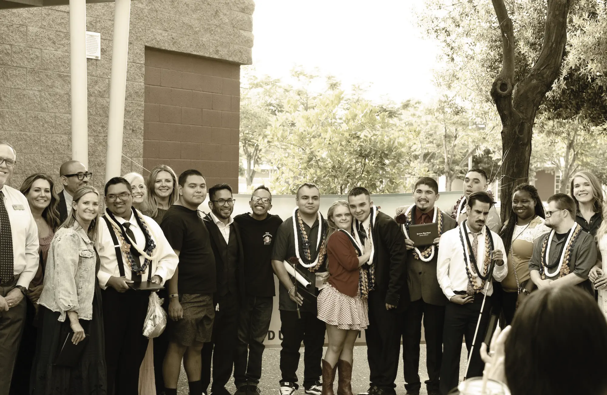 Group photo of young adults with moderate to severe disabilities and staff from the BRIDGE program, standing outdoors after a graduation ceremony. Many of the young adults are wearing celebratory leis.