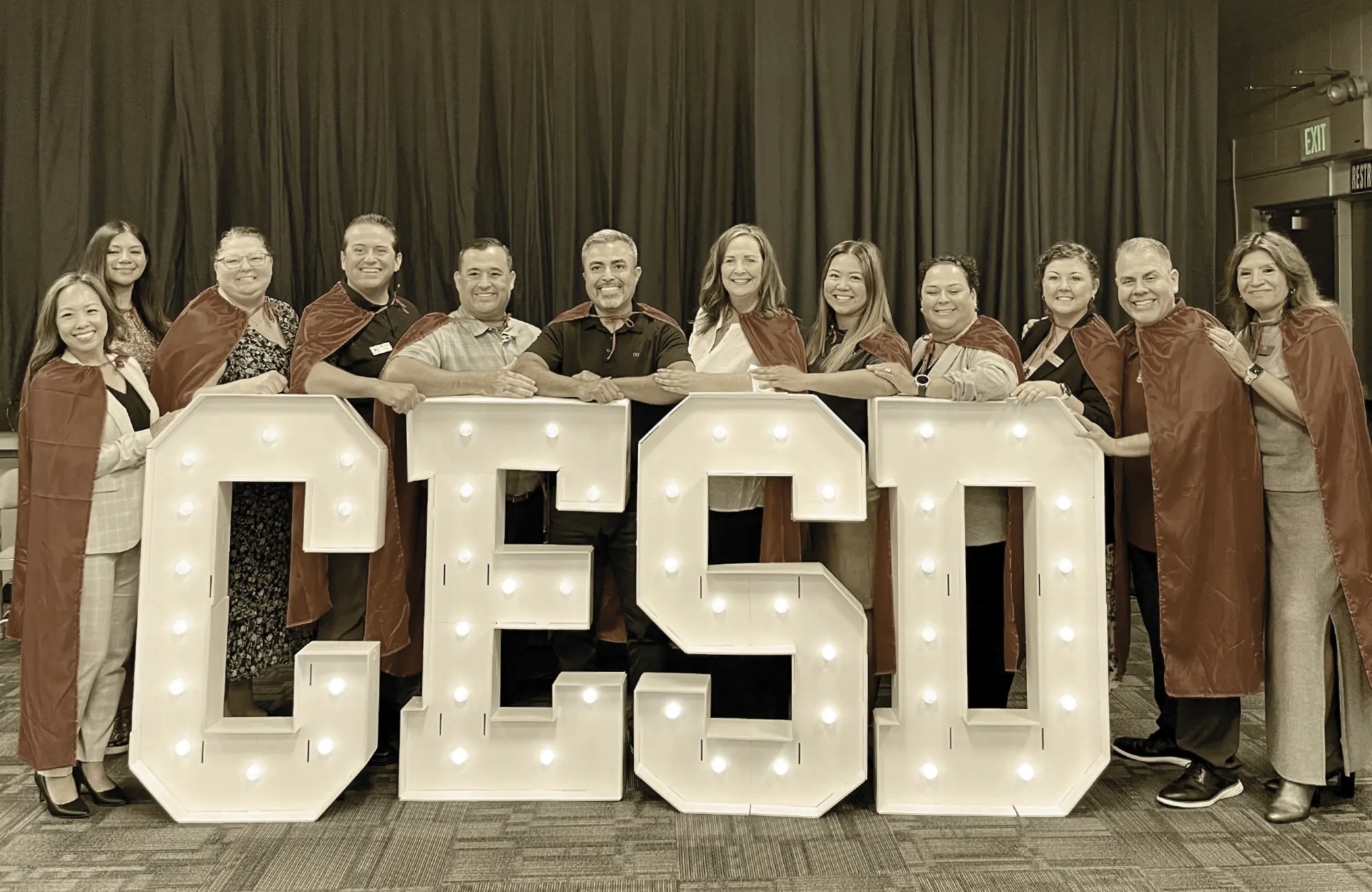 Group of twelve smiling Centralia ESD staff members wearing red superhero capes and posing behind large, illuminated marquee letters that spell out "CESD."