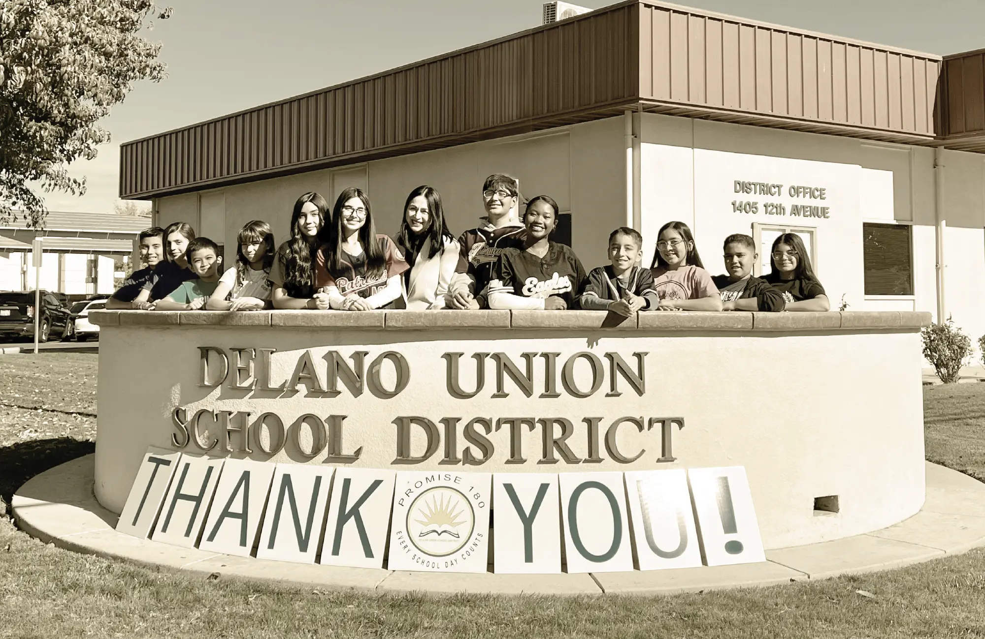 Twelve smiling middle school students and an adult pose behind a curved Delano Union School District sign. Large letters spelling "THANK YOU" are propped in front of the sign.