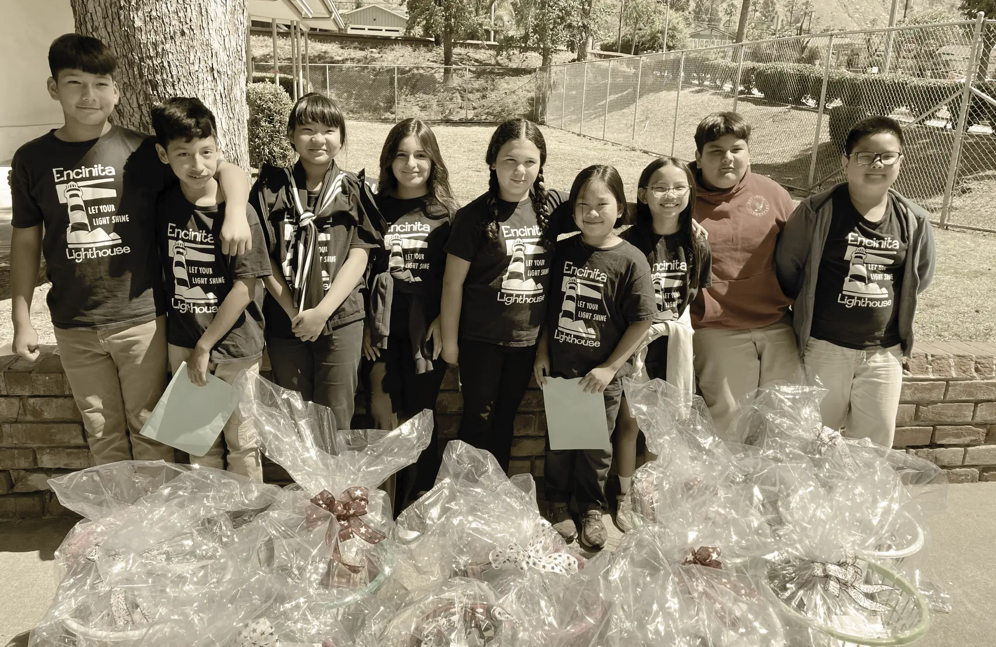 Nine elementary school students wearing "Encinitas Lighthouse" shirts stand outdoors behind multiple wrapped gift baskets they helped prepare as part of a service project.