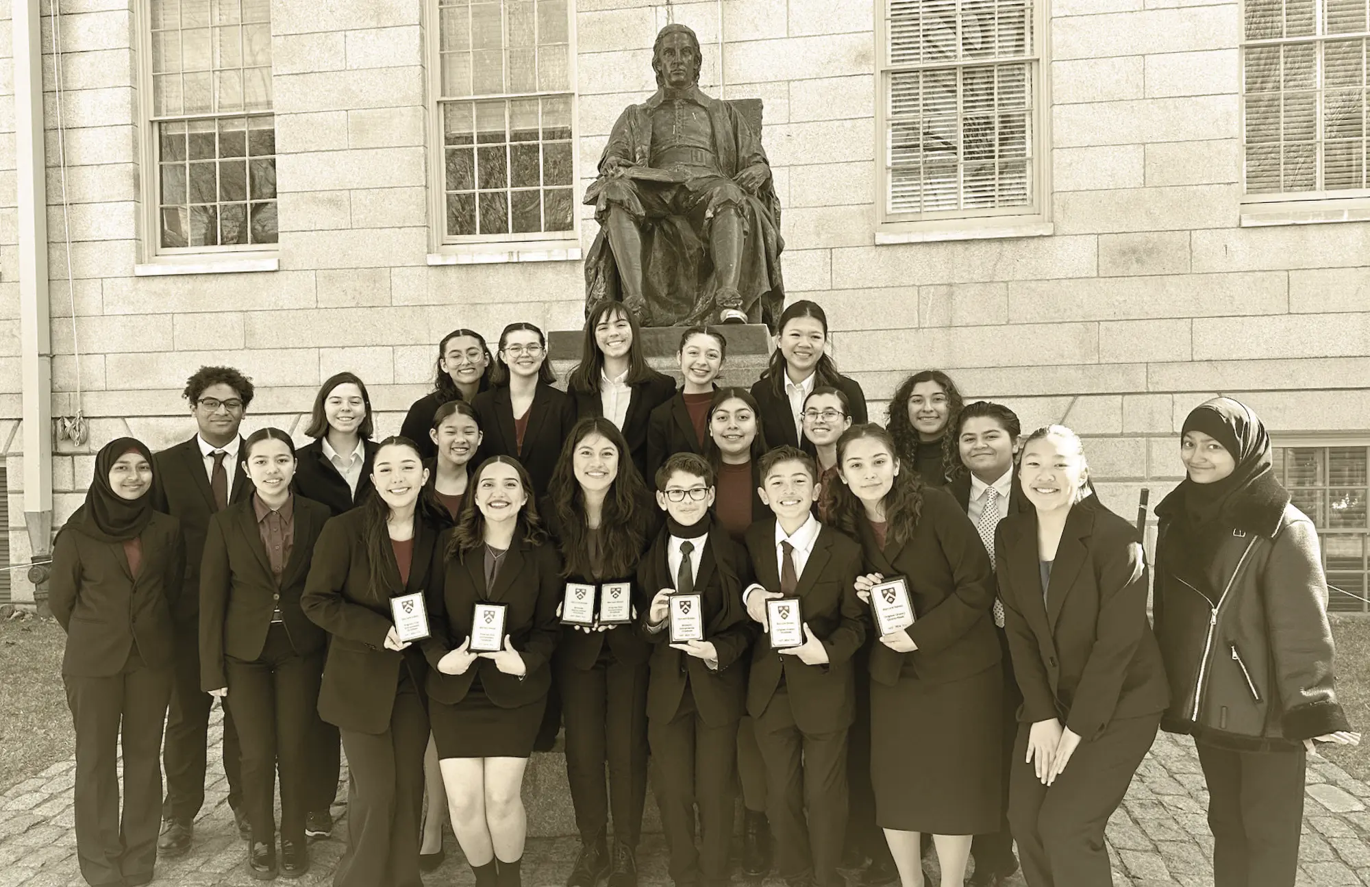 Group photo of a high school Speech and Debate team standing in front of a large stone building and statue. Several students in the front row are holding trophies and awards.