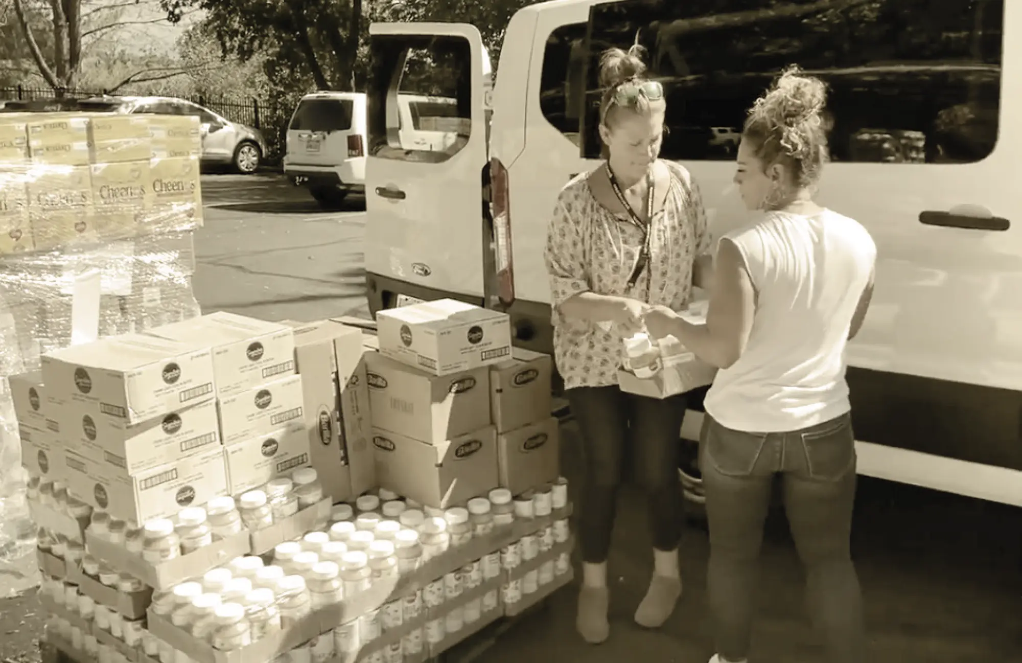 Two women loading boxes of food items and large pallets of shelf-stable goods, including jars and cereal boxes, from a parking lot into a white cargo van for the food pantry program.