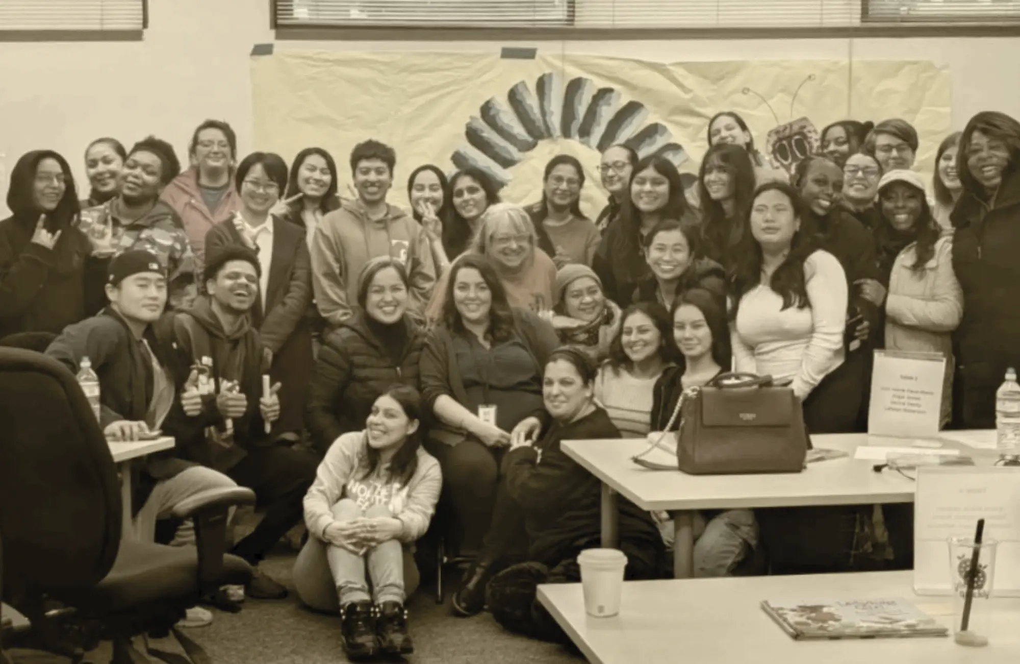 A large, diverse group of adults, members of an education workforce cohort, smile while gathered in a classroom setting in front of a painted mural background.