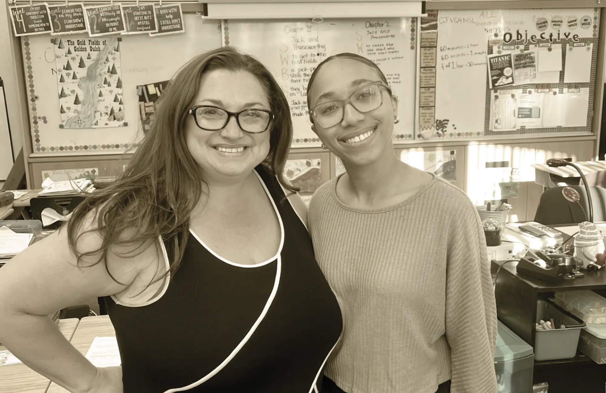 Two women, one a veteran teacher and the other a younger resident, smile together in a brightly lit classroom, representing the success of a teacher pipeline program.