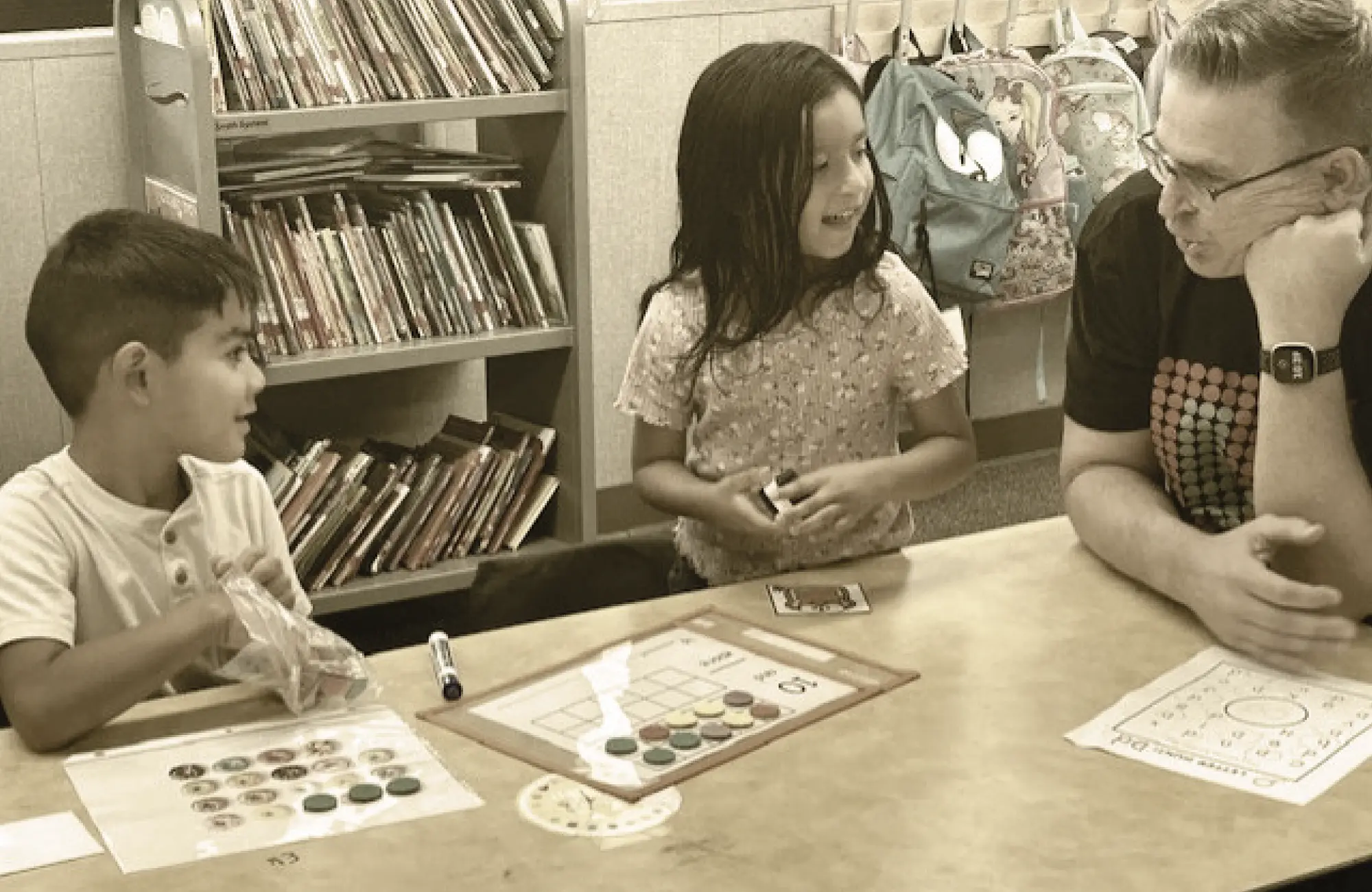 Two young students engage in a hands-on math and literacy intervention activity at a table with an adult tutor or teacher. Learning materials are spread out in front of them.
