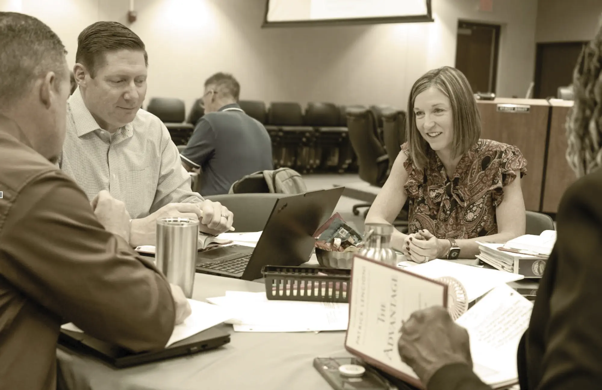 Four educational leaders are seated around a table for a meeting, focused on a laptop and documents. This represents the daily, data-driven collaboration of the Daily Huddle Program.