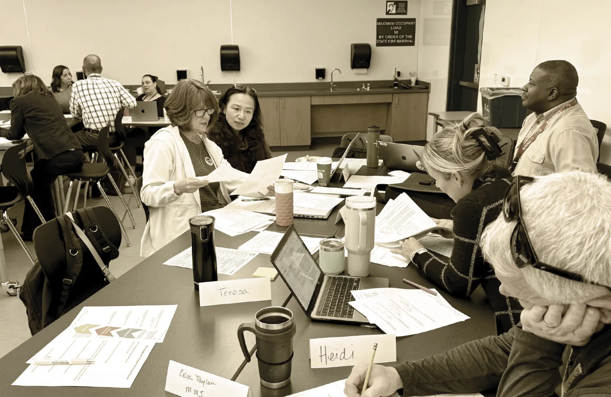 Teachers collaborate in a professional development setting, seated around a table covered in data sheets, notes, and laptops, representing a data-driven protocol meeting.