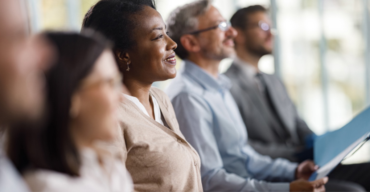 A smiling woman sits in a row with other professionals, focusing on a presentation during a board development seminar.