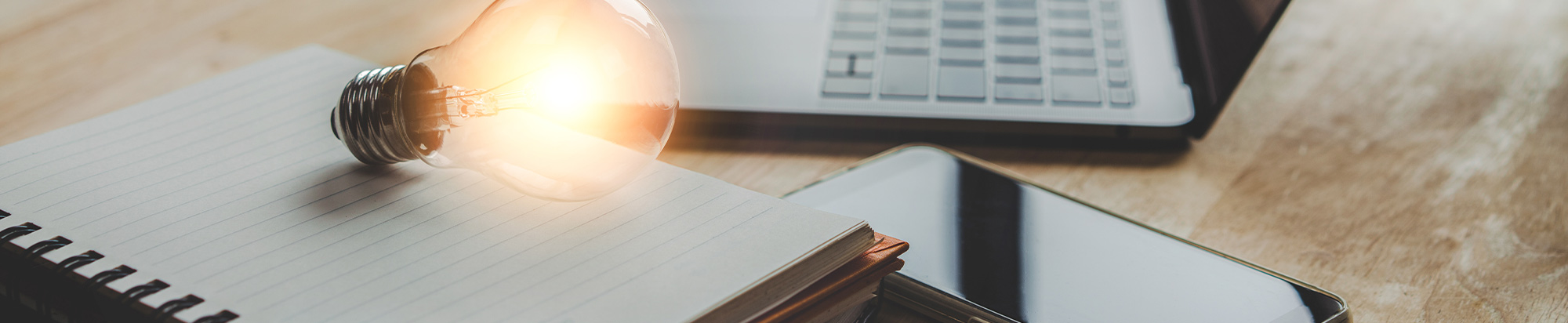 A glowing incandescent lightbulb resting on a spiral notebook next to a laptop and smartphone.