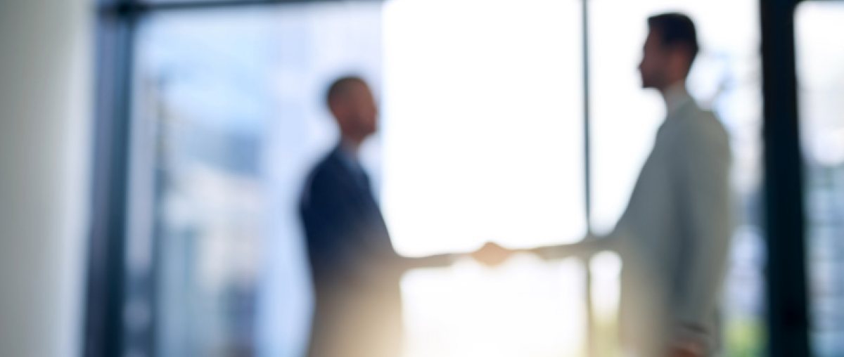 A blurred photograph silhouette of two male professionals shaking hands in front of a large, bright window in a modern office building