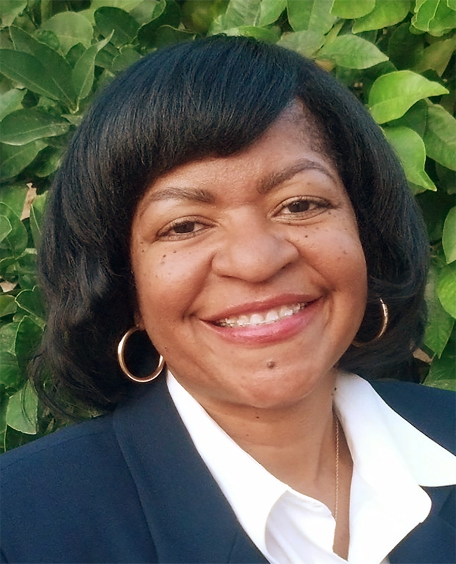 Close-up headshot of Angelina Pride, a smiling woman with dark, chin-length hair styled in a bob with bangs; She is wearing gold hoop earrings, a white collared shirt, and a navy-blue blazer, posing against a background of green leafy foliage