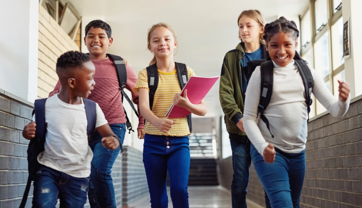 A candid shot of five diverse elementary-aged children smiling and running down a bright, sunlit school hallway while wearing backpacks