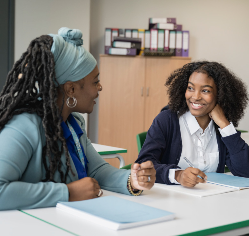 A young woman in a classroom smiles while looking at an instructor wearing a blue headwrap during a discussion.