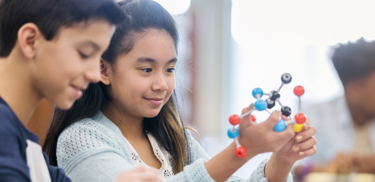A young student carefully holds and examines a colorful molecular model while working with a classmate in a science lab.