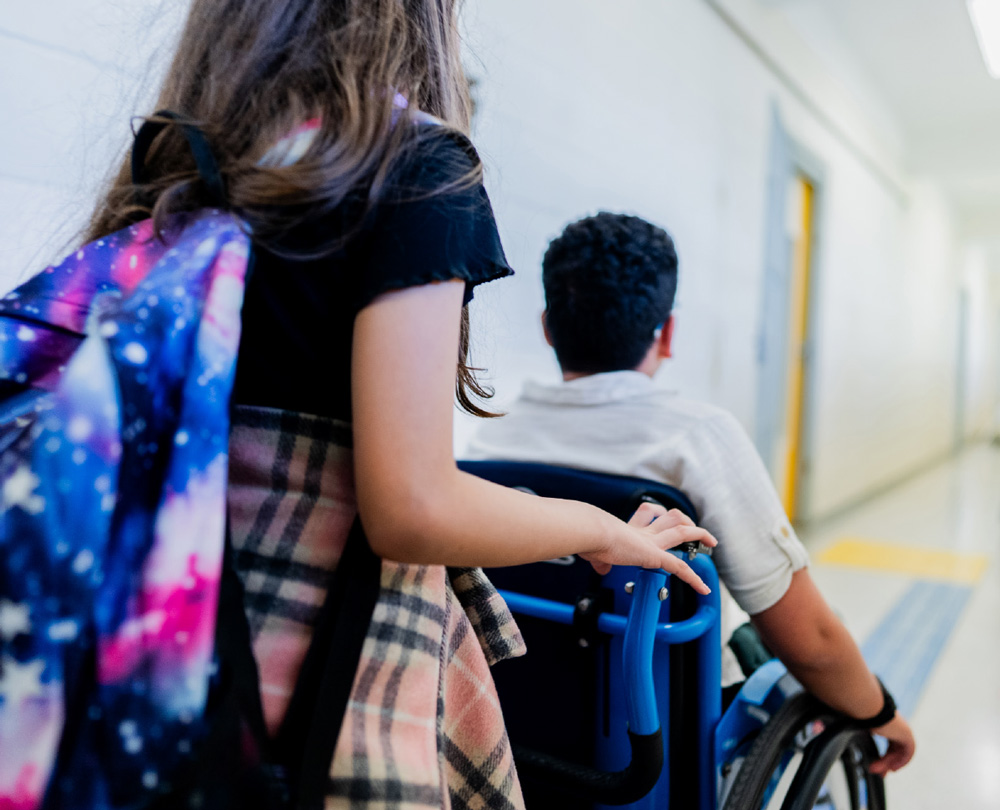 A student with a galaxy-print backpack follows behind a classmate using a blue wheelchair down a school hallway.