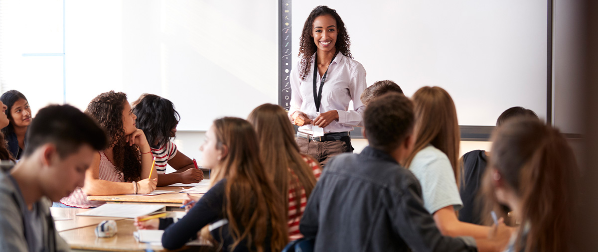 A smiling female teacher standing at the front of a diverse high school classroom while students listen and take notes at their desks.