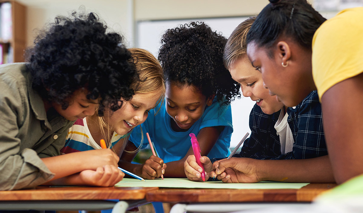Five diverse students lean over a wooden desk, drawing and writing on a large sheet of green paper with colorful pens and pencils.