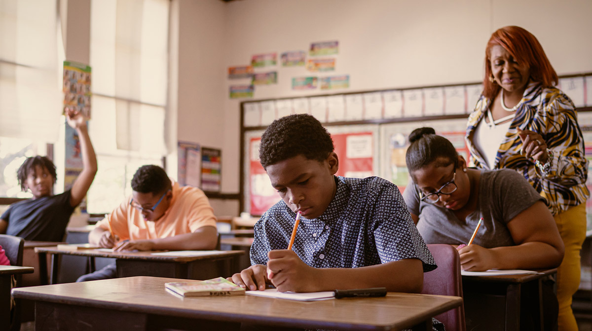 Students sitting at wooden desks in a sunny classroom, diligently working on an assignment as their teacher in a patterned blazer observes from the background.