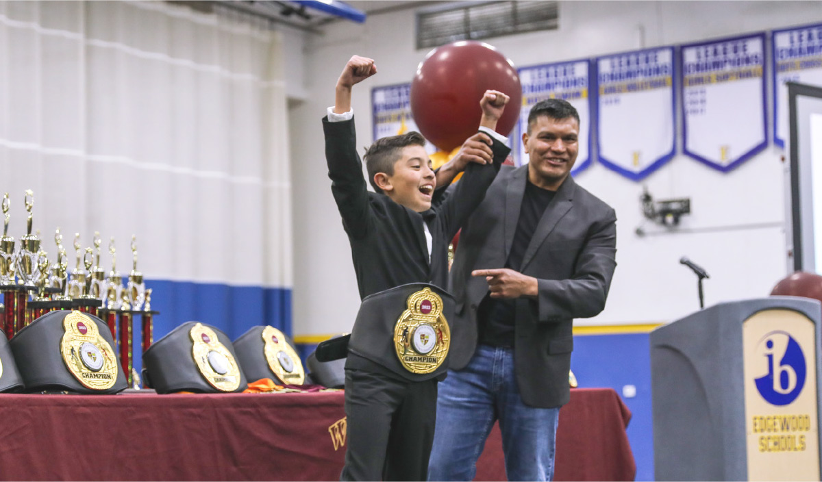 A young boy in a suit raises his fists in victory while wearing a champion belt, standing next to a smiling man in a gym.