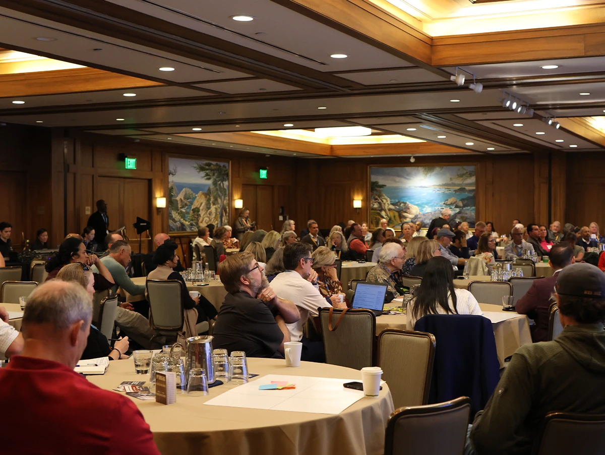 A large group of professionals attending a seminar or workshop in a wood-paneled conference room with round tables.