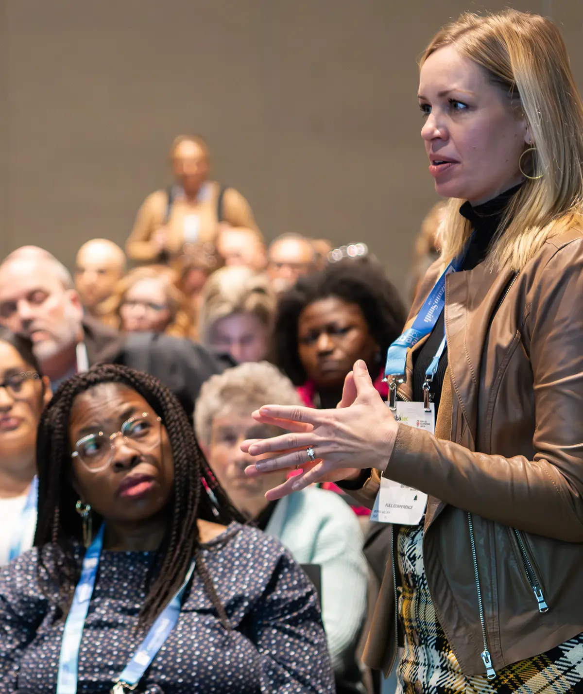 A female attendee in a brown leather jacket and plaid skirt asking a question during a member services Q&A session at a professional conference.