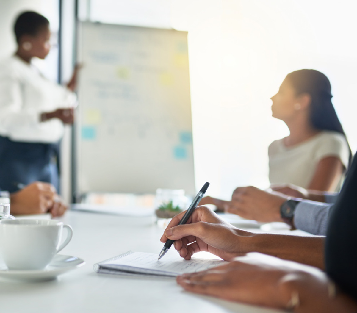 A professional attending a board governance workshop, taking detailed notes while a presenter stands at a whiteboard in the background.