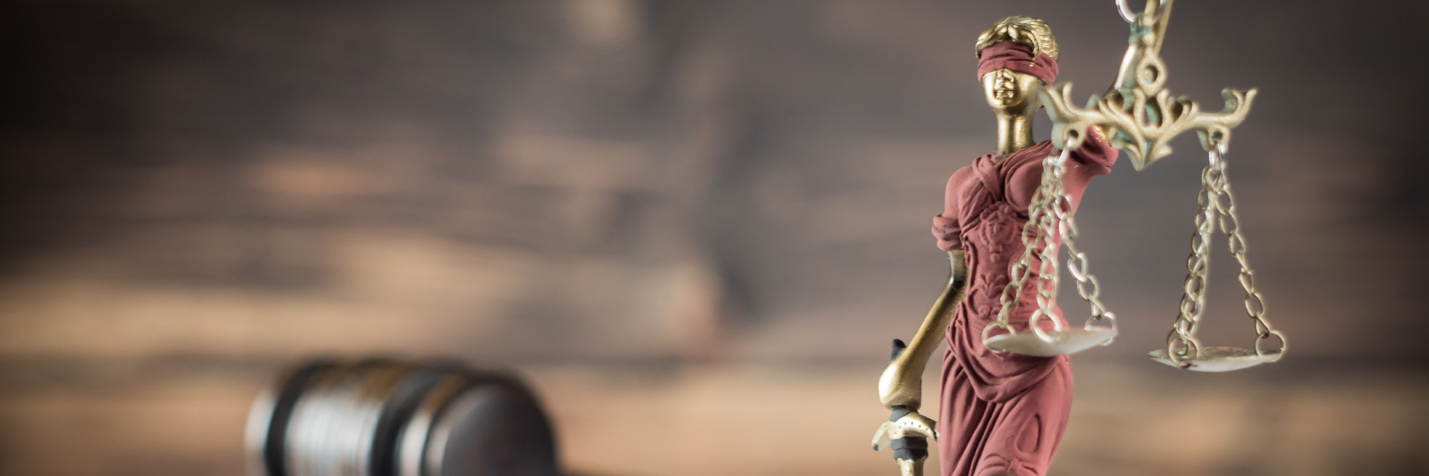 A gold and bronze Lady Justice statue holding scales stands in front of a blurred wooden background with a gavel.