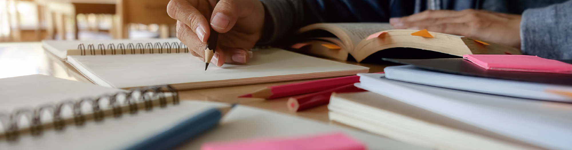 A close-up view of a student's hand using a pencil to write in a notebook, surrounded by textbooks and colorful sticky notes.