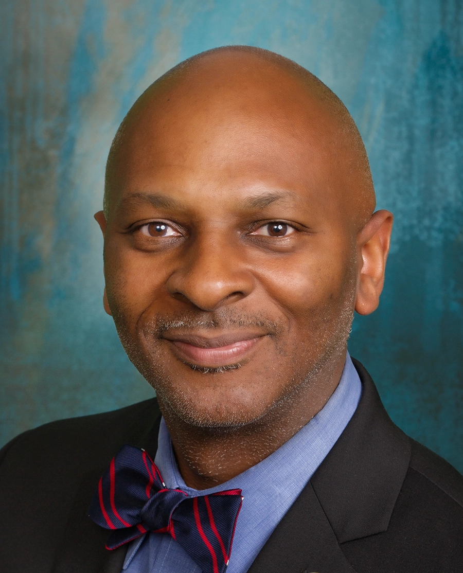 Close-up headshot of Daryl Camp, a smiling man with a shaved head; He is wearing a blue dress shirt, a navy-blue and red striped bowtie, and a black suit jacket against a textured blue-green background