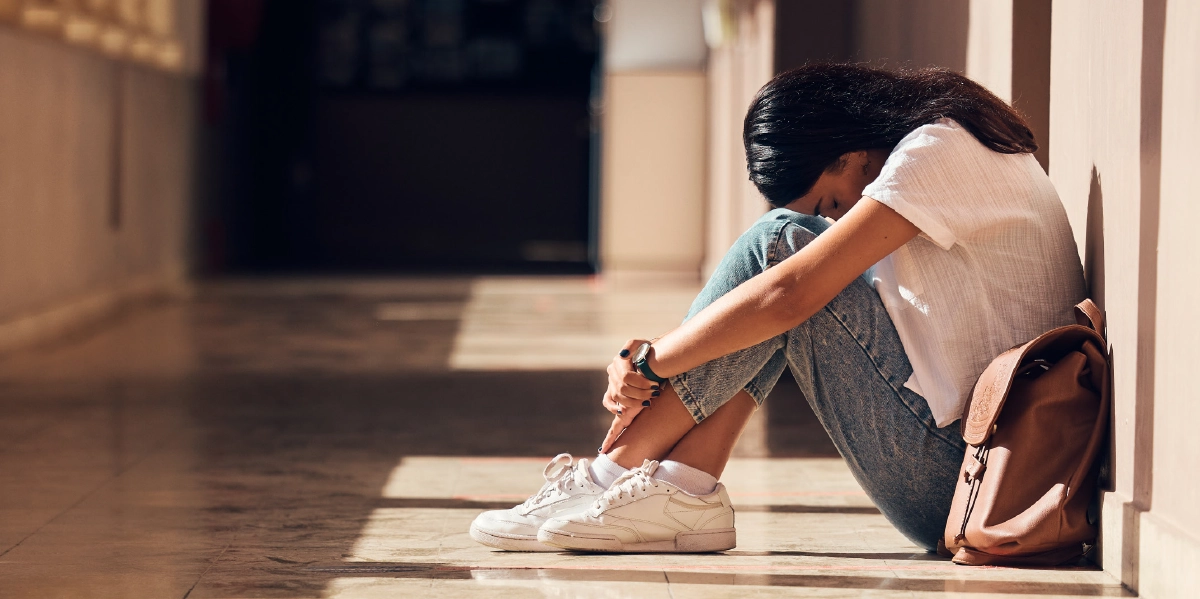 A young female student sits on the floor of a sunlit school hallway, leaning against the wall with her head resting on her knees in a dejected or weary pose; Her backpack sits beside her on the polished floor, which reflects the light from the windows