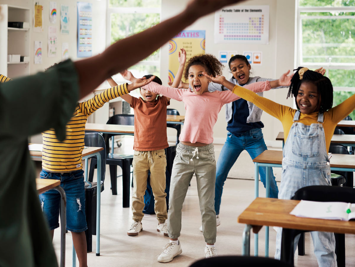 A diverse group of young children in a classroom joyfully participating in an activity with their arms raised and mouths open as if cheering or singing.