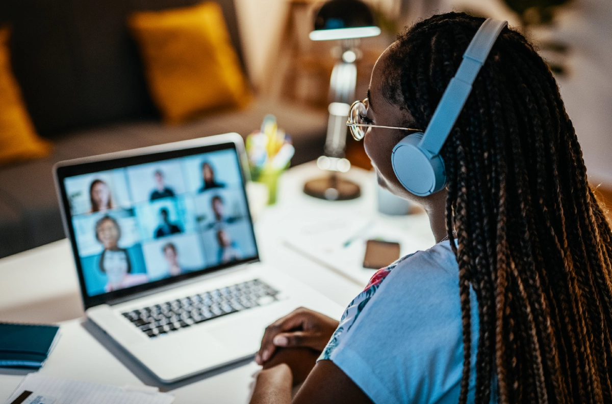 A high-angle, over-the-shoulder shot of a female person with long braids wearing blue headphones and glasses while participating in a video conference on a laptop; The screen shows a grid of several other participants, and the person is sitting at a white desk in a dimly lit, cozy room