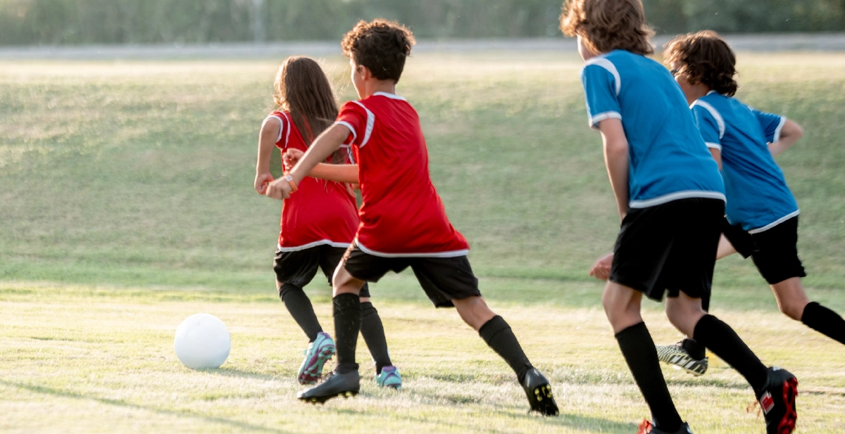 An action shot from a youth soccer game on a grassy field; Four children in red and blue jerseys are seen from behind, running toward a white soccer ball in the late afternoon sun