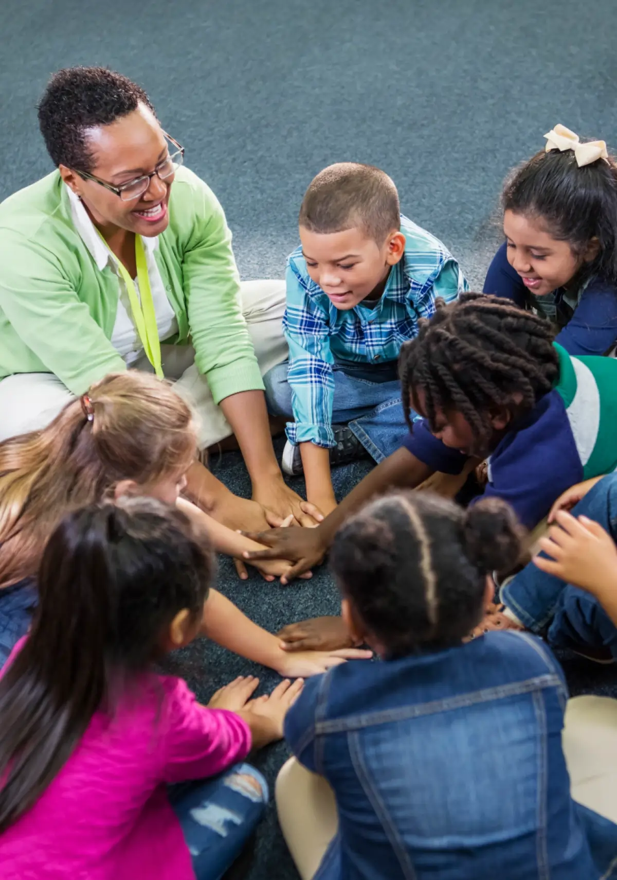 A smiling female teacher with short dark hair and prescription glasses sitting on a carpet with a diverse group of young kid students